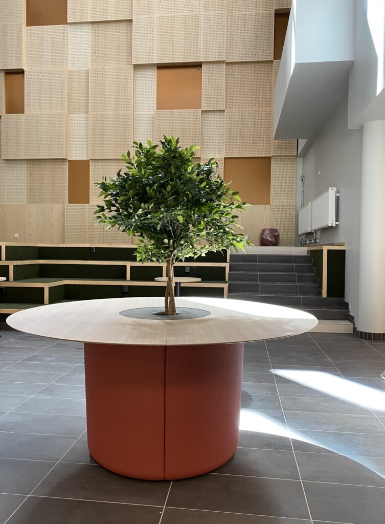 Konstgjort träd i skolmiljö placerat i sittmöbel med integrerad växtinredning i gemensam yta